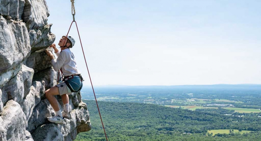 A person wearing safety gear is secured by ropes as they rock climb high above a vast forested area below. 